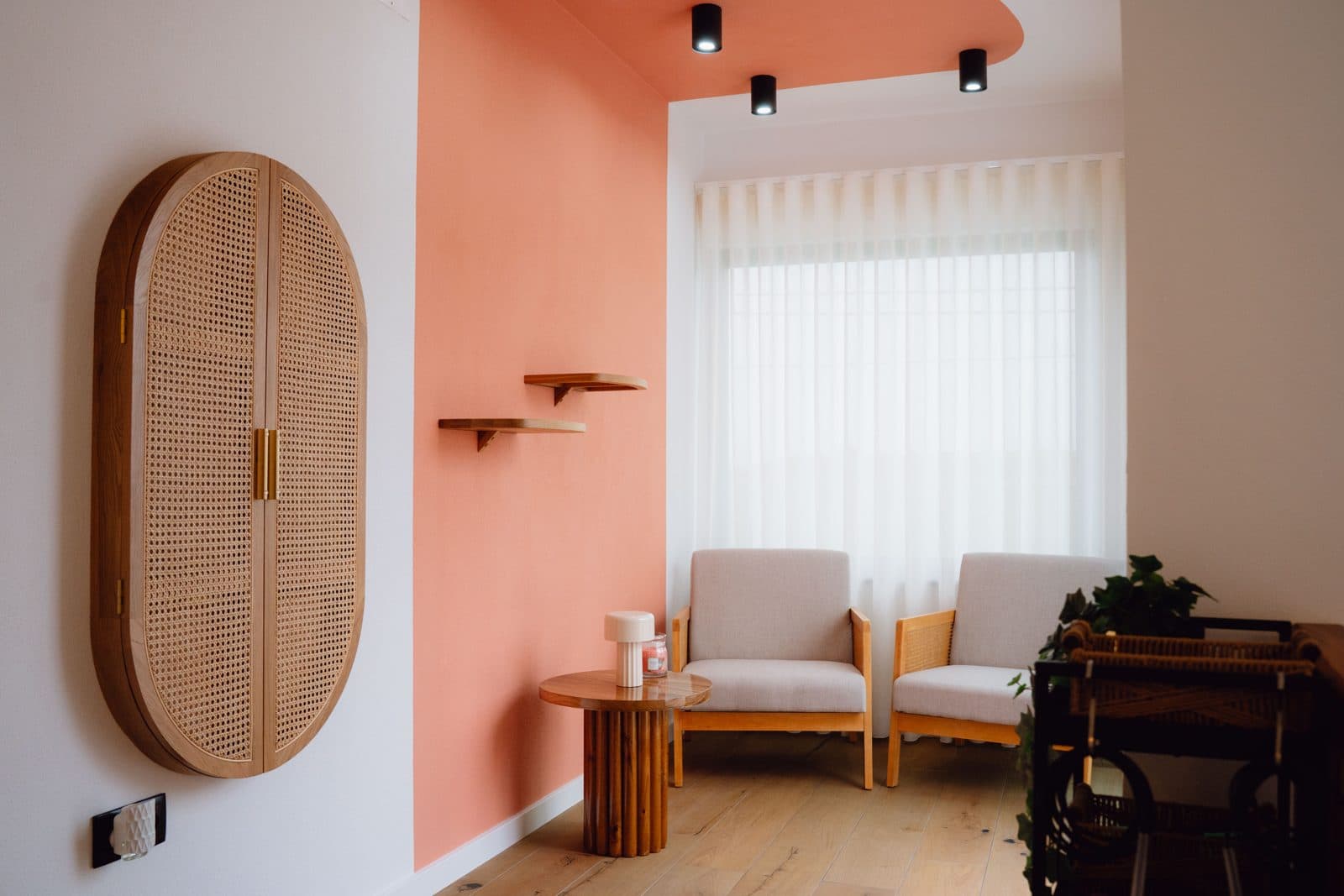 Canto de leitura do Terrassi Villa: duas poltronas claras de estrutura em madeira junto a uma janela com cortinado transparente, mesa de apoio redonda com candeeiro, prateleiras de madeira numa parede coral e um armário oval de palhinha — vista 1