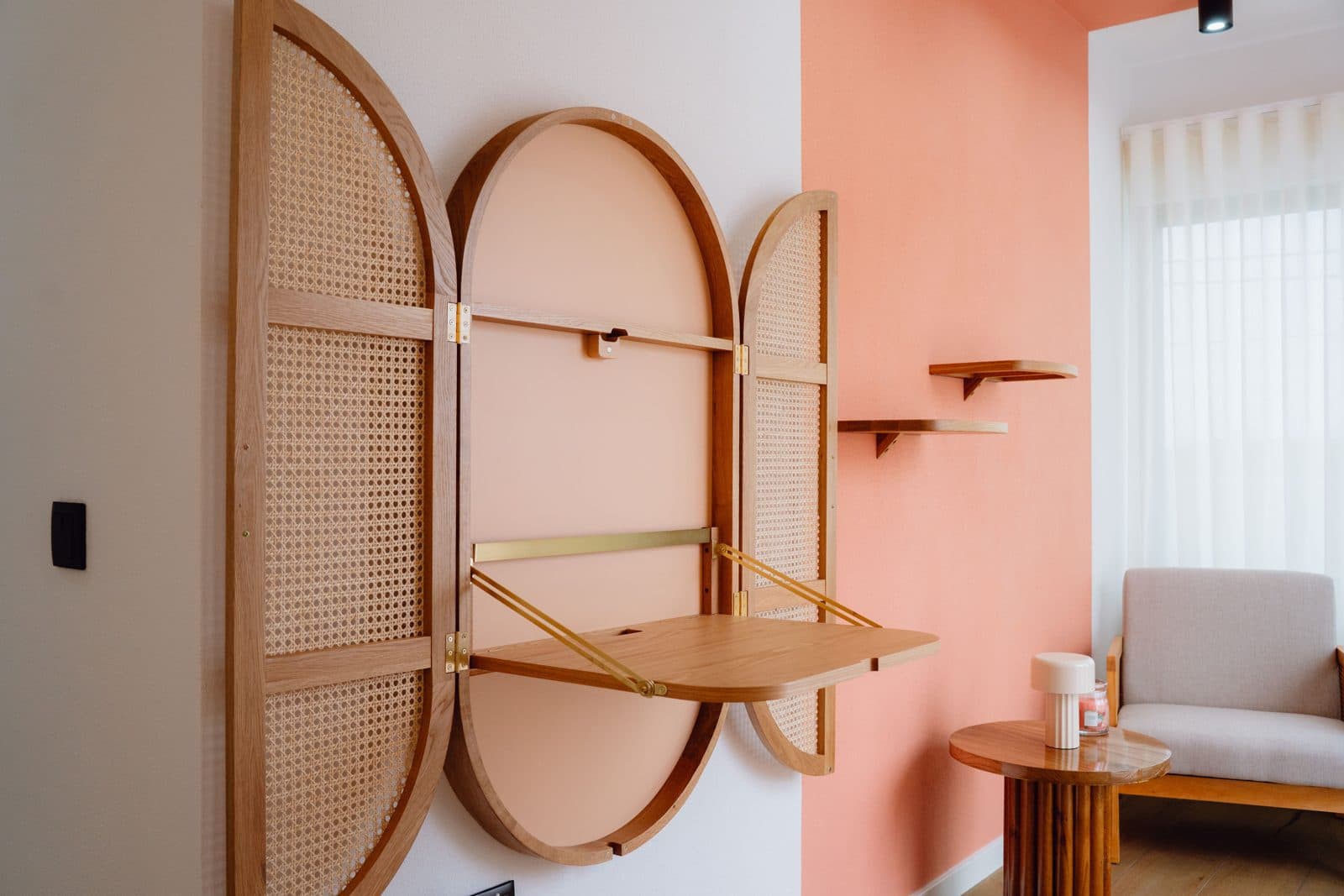 Canto de leitura do Terrassi Villa: duas poltronas claras de estrutura em madeira junto a uma janela com cortinado transparente, mesa de apoio redonda com candeeiro, prateleiras de madeira numa parede coral e um armário oval de palhinha — vista 2