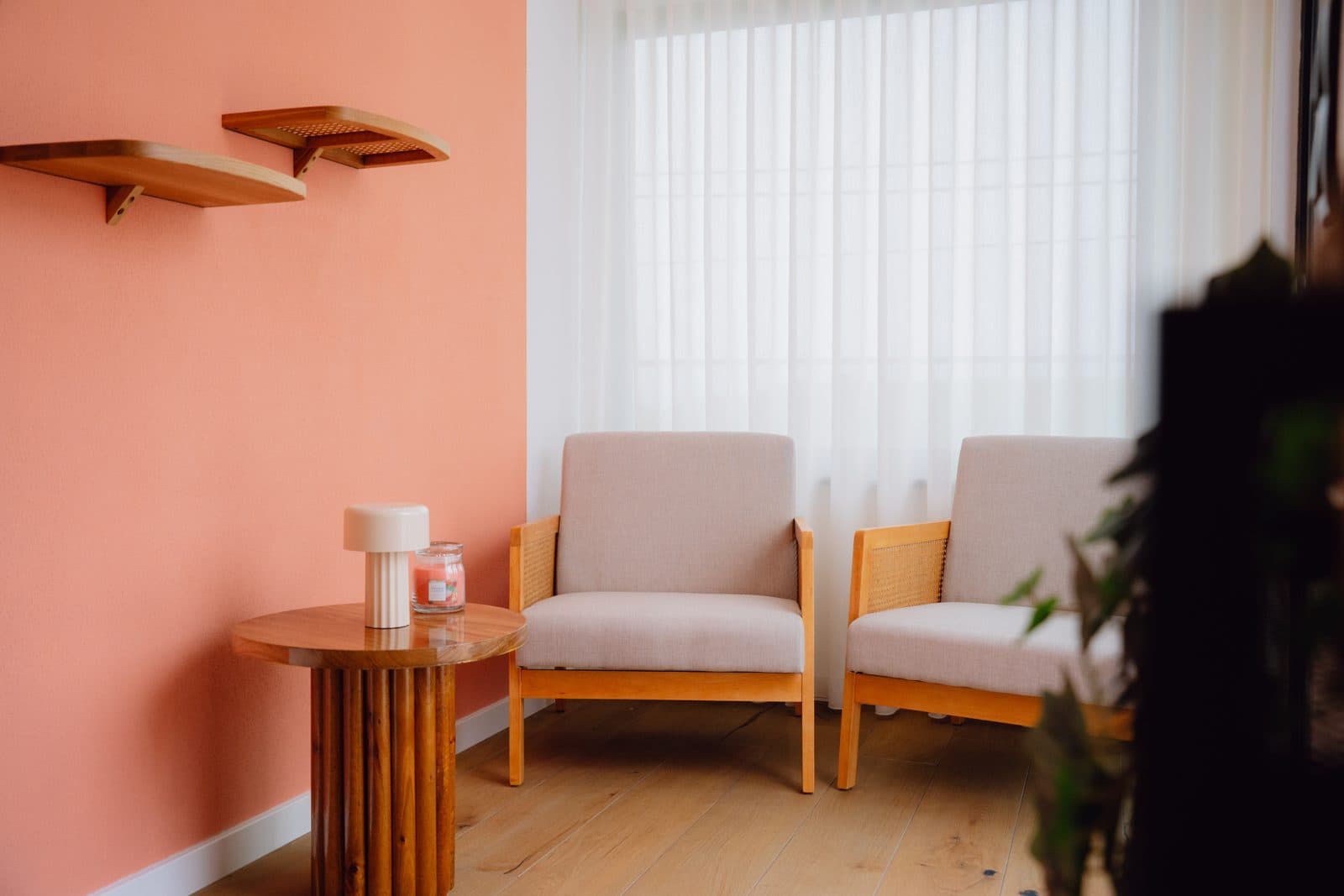 Canto de leitura do Terrassi Villa: duas poltronas claras de estrutura em madeira junto a uma janela com cortinado transparente, mesa de apoio redonda com candeeiro, prateleiras de madeira numa parede coral e um armário oval de palhinha — vista 3
