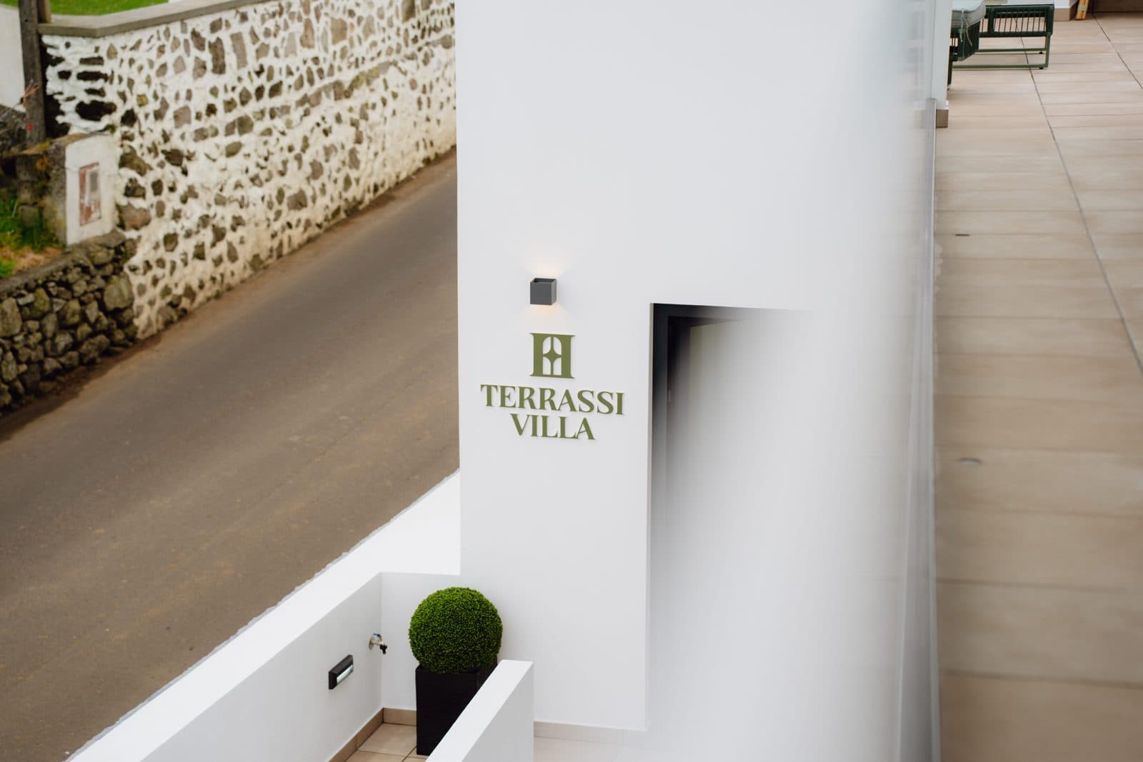 Exterior do Terrassi Villa: volumes brancos com a placa da casa em verde, terraço pavimentado com sofá e porta de correr para o interior, e em redor o pasto e os muros de pedra basáltica da costa norte da Terceira — vista 2