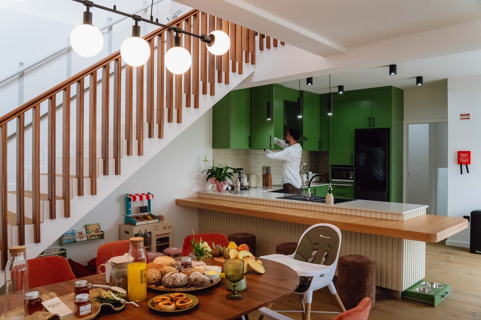Open space do Terrassi Villa: cozinha verde com bancada corrida em madeira, mesa de refeição posta e, sob a escada de ripado, um recanto de brincar com banca de mercado, cozinha de madeira e livros infantis — vista 1