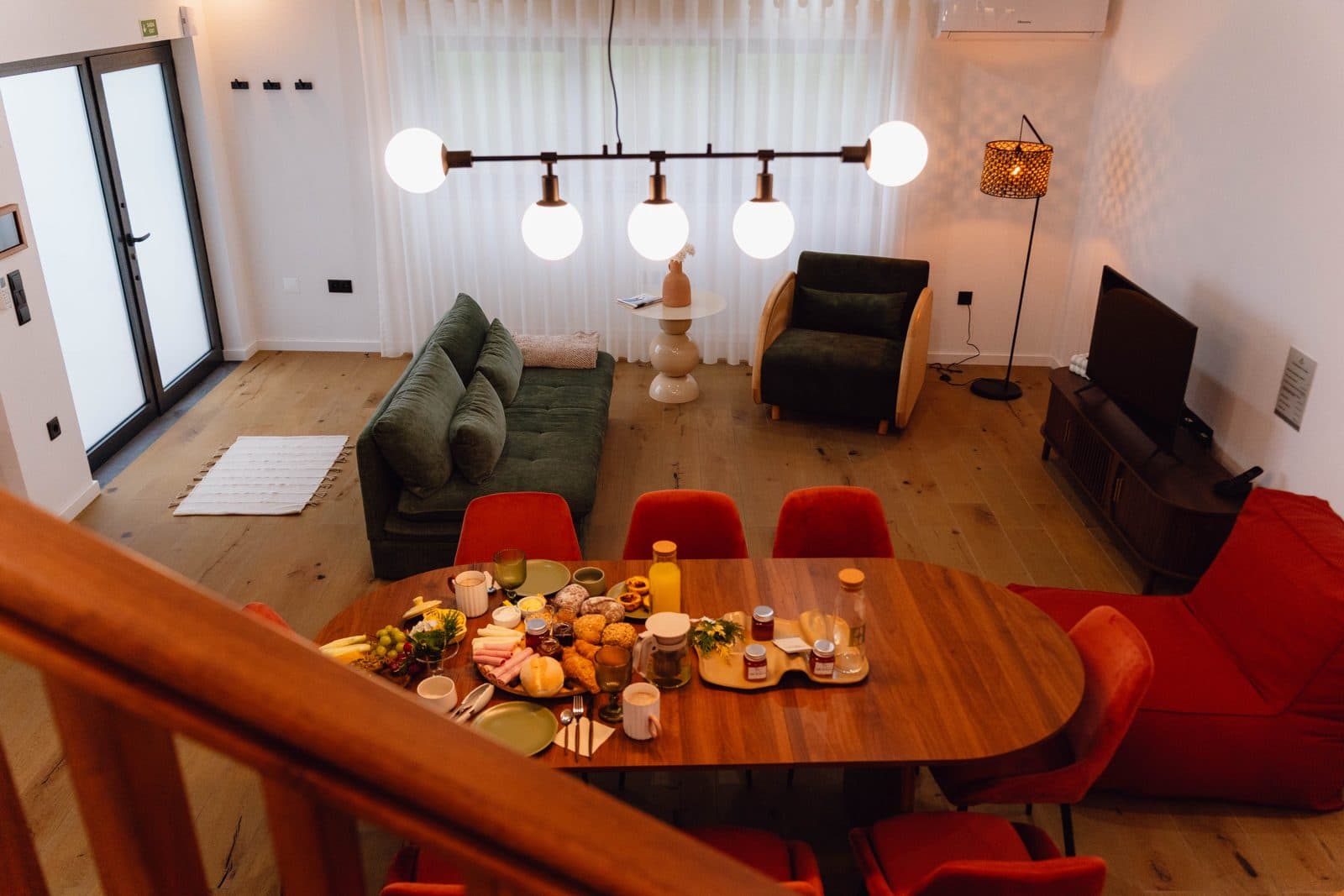 Open space do Terrassi Villa: cozinha verde com bancada corrida em madeira, mesa de refeição posta e, sob a escada de ripado, um recanto de brincar com banca de mercado, cozinha de madeira e livros infantis — vista 2