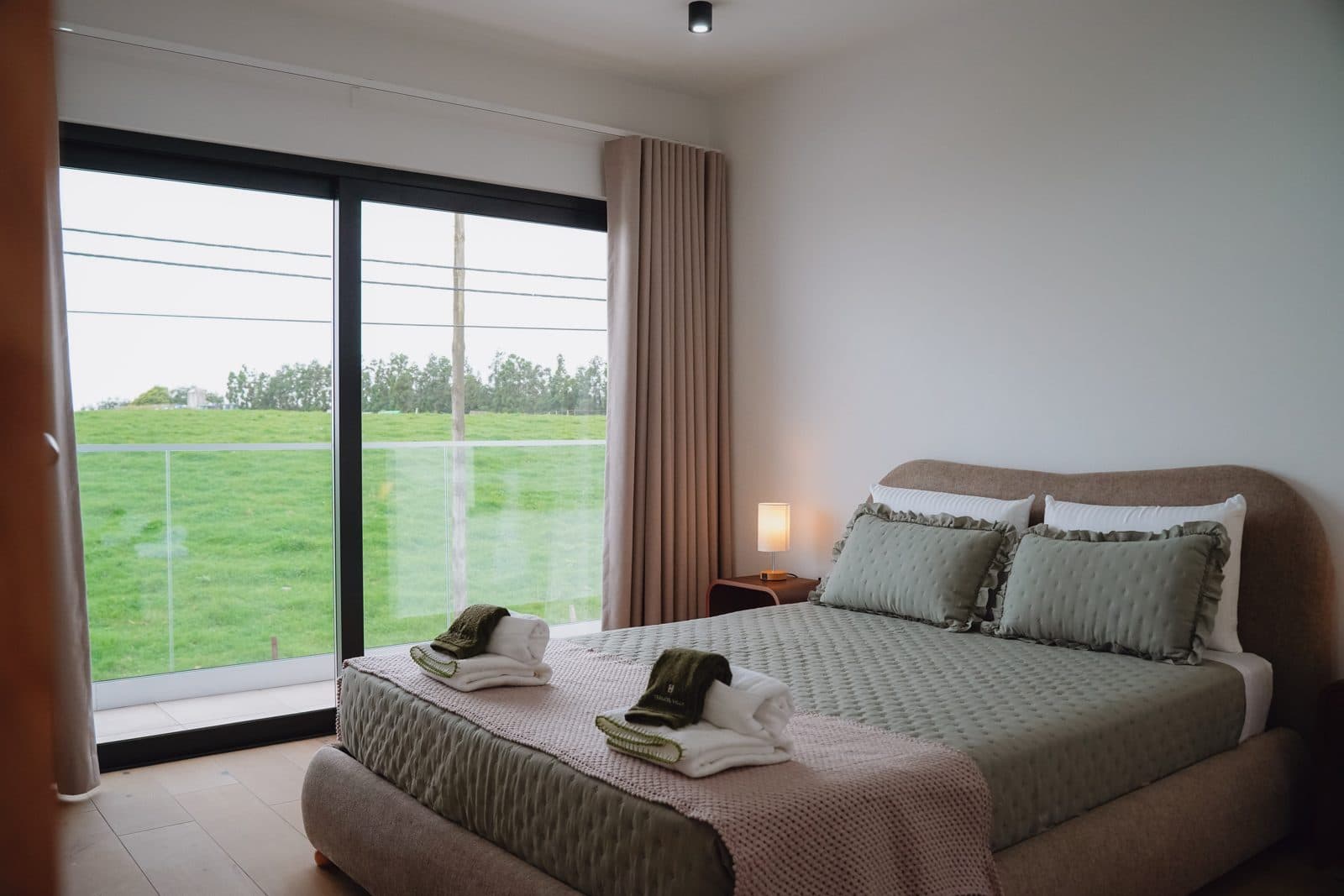 Quarto do Terrassi Villa: cama de casal com colcha verde-seiva e almofadas de folho, cabeceira estofada, mesinhas suspensas em madeira com candeeiro aceso, e porta de correr para uma varanda com guarda de vidro sobre o pasto — vista 2