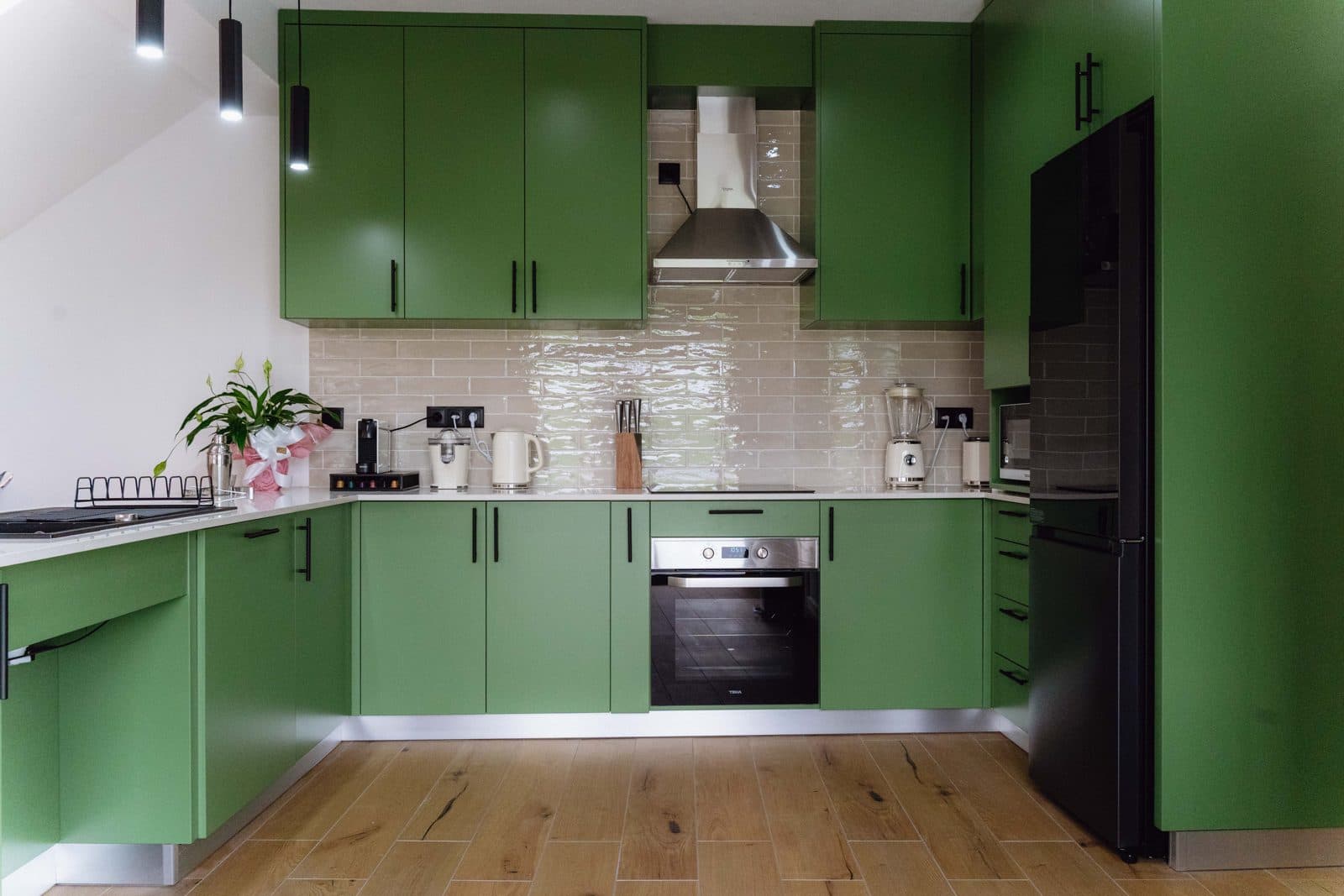 Cozinha adaptada do Terrassi Villa: armários verdes em U, azulejo bege brilhante, forno encastrado, placa de indução e exaustor; à esquerda, um troço de bancada com espaço livre por baixo, e uma gaveta funda aberta com tachos ao alcance da mão — vista 1