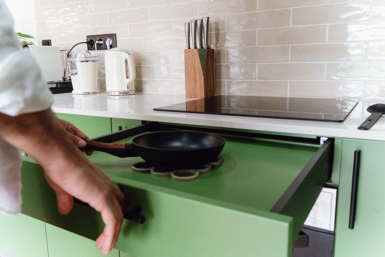 Cozinha adaptada do Terrassi Villa: armários verdes em U, azulejo bege brilhante, forno encastrado, placa de indução e exaustor; à esquerda, um troço de bancada com espaço livre por baixo, e uma gaveta funda aberta com tachos ao alcance da mão — vista 3