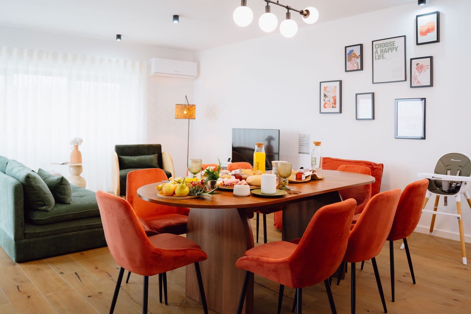Sala e sala de jantar integradas do Terrassi Villa: mesa de madeira posta com cadeiras em veludo terracota, sofá e poltrona verdes junto à janela, televisão sobre móvel de madeira, parede com molduras e chão livre entre as 3 áreas — vista 1
