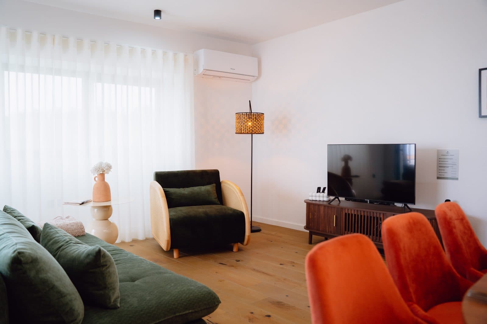 Sala e sala de jantar integradas do Terrassi Villa: mesa de madeira posta com cadeiras em veludo terracota, sofá e poltrona verdes junto à janela, televisão sobre móvel de madeira, parede com molduras e chão livre entre as 3 áreas — vista 2
