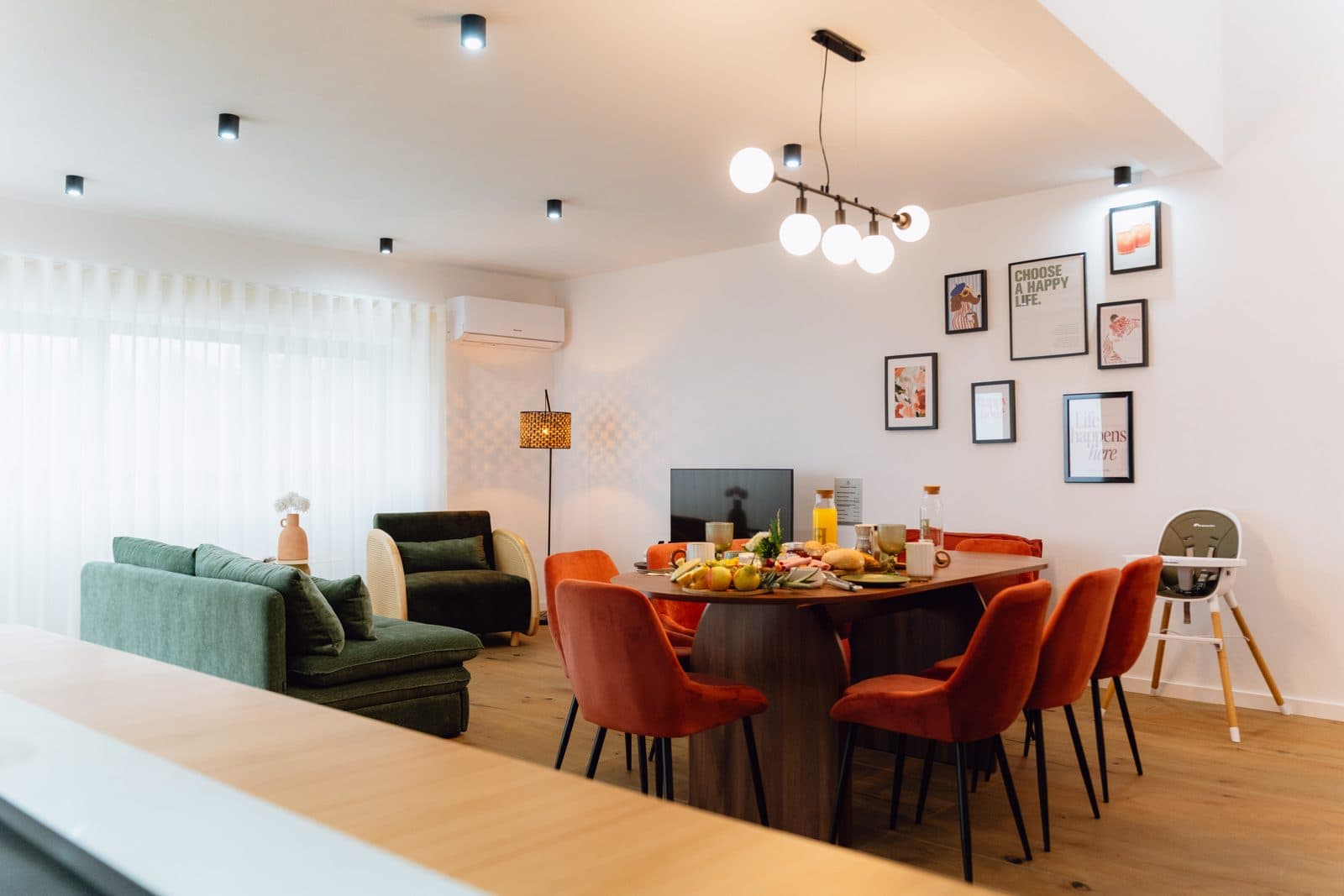 Sala e sala de jantar integradas do Terrassi Villa: mesa de madeira posta com cadeiras em veludo terracota, sofá e poltrona verdes junto à janela, televisão sobre móvel de madeira, parede com molduras e chão livre entre as 3 áreas — vista 3