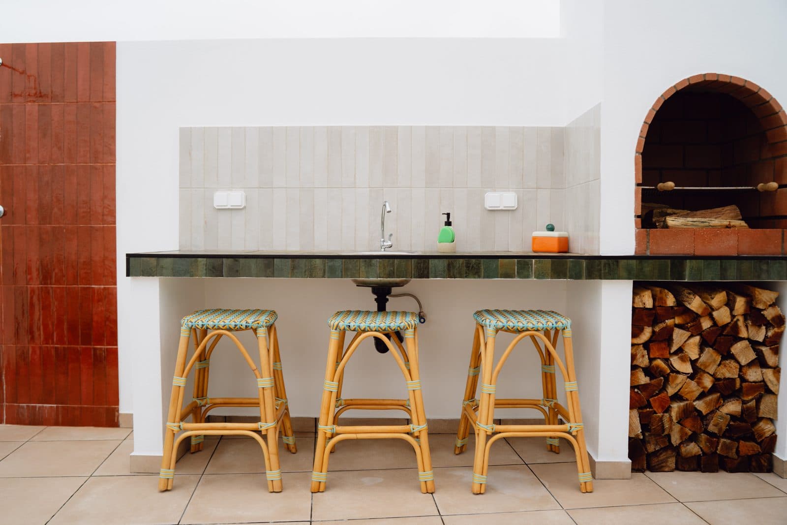 Terraço do Terrassi Villa: bancada exterior com lava-loiça e remate em azulejo verde, três bancos altos de verga, e forno de lenha em tijolo com arco e lenha empilhada por baixo da bancada — vista 1
