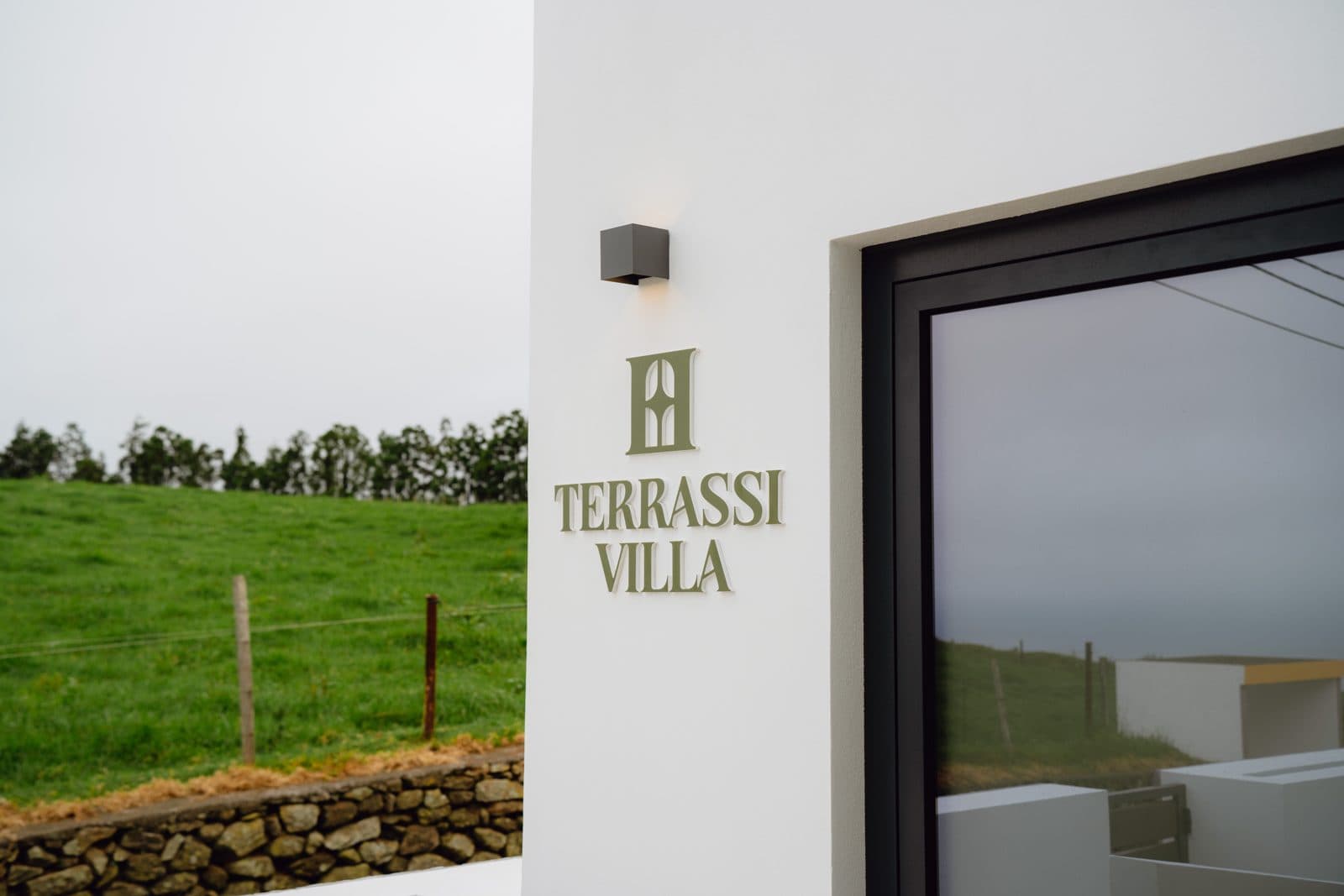 Exterior do Terrassi Villa: volumes brancos com a placa da casa em verde, terraço pavimentado com sofá e porta de correr para o interior, e em redor o pasto e os muros de pedra basáltica da costa norte da Terceira