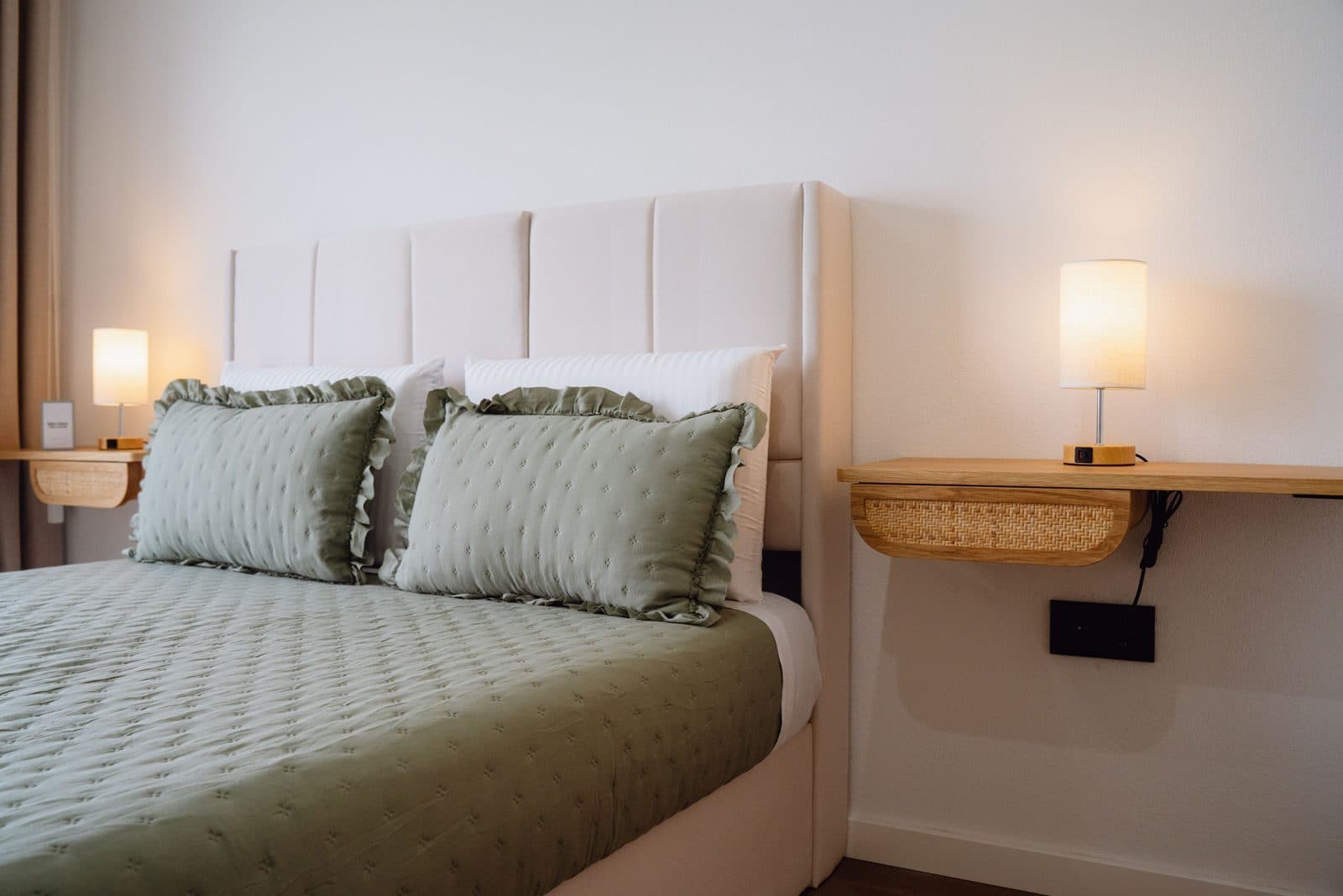 Quarto do Terrassi Villa: cama de casal com colcha verde-seiva e almofadas de folho, cabeceira estofada, mesinhas suspensas em madeira com candeeiro aceso, e porta de correr para uma varanda com guarda de vidro sobre o pasto